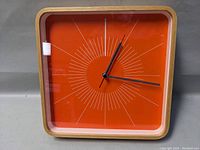 Front view of square orange clock face with radiating white line design and black hands in wood frame.