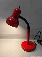 Red gooseneck desk lamp with curved shade, flexible arm and round base, shown against plain background, illuminated to show working bulb