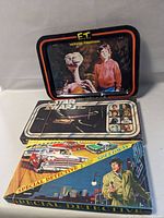 Photo of the E.T. folding tray at the back standing upright, with Star Wars and Special Detective Speedway board games stacked in front with rubber bands around them.