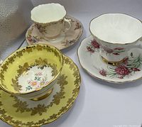 Three vintage teacups with matching saucers showing distinct floral patterns and gold trim.