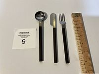 Three-piece flatware set laid out with ruler for scale, showing spoon, knife, and fork with dark gray handles and shiny metal ends.
