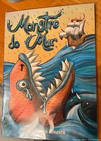 Close-up of the front cover of the Monstro do Mar moving paper theatre, showing a stylized orange sea monster with large teeth and tear drops, with a sailing ship in the ocean.