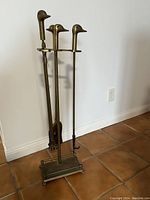 Full view of the mallard head brass fireplace tool set with stand against a white wall on a tiled floor.