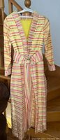 Front view of a long silk dress with yellow, pink, and white horizontal stripes, featuring a V-neck with ruffles, long sleeves, and a matching fabric belt tied at the waist.