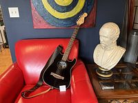 Black Fender Stratacoustic guitar with leather strap shown on red chair next to non-included bust and painting