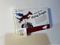 Front of the box showing the assembled wooden dragon automaton on a base with flying pose and movement wires, factory sealed packaging with product name and age recommendation.