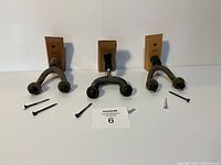 Three guitar hangers arranged side by side on a white surface, each with a wooden mounting plate and black padded arms. Assorted mounting screws are displayed in front of the hangers.