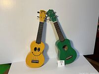 Pair of two ukuleles, one yellow with a smiley face and one green, standing against a white background.