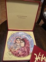 Porcelain collectors plate inside original hinged presentation box, showing detailed painted image of mother and child by Edna Hibel, gold gilded edges visible.