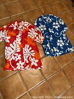 Photo of two vintage men's Hawaiian shirts laid on tile floor. Orange shirt with white and orange floral pattern on the left, blue shirt with white floral print on right.