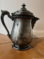 Front view of antique silver plate coffee pot water jug showing floral engraving, curved handle, lid with finial, and patina/dents.