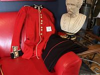 Full view of the red British Soldiers tunic and black trousers laid on a red couch with a white bust statue nearby.