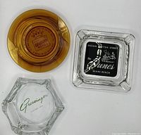 Top-down view of three vintage ash trays including amber King Sleep-Well, square 'the Dunes Miami Beach', and hexagonal Grossingers ash trays.