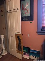 Wall with closed door displaying a framed Erte poster and hanging fabric rubbing with some paintings leaning against the wall below