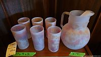 Front view of six frosted glass tumblers and one pitcher with mottled pink and peach decoration displayed on a wooden shelf.