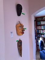 Three masks hanging vertically on a wall; dark wood mask at top, yellow/red face mask in middle, elongated earthy tone mask at bottom.