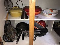 Shelf with yellow and red Dutch ovens, cast iron skillets, metal funnel, and various kitchen utensils including strainers and baskets.