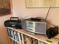 Panasonic black boombox and Sony ZS-D5 audio system with remote placed on a shelf above books, showing front and top views with antenna extended.