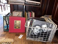 Wide photo showing wire basket full of various vinyl record albums with visible covers including a black and white photo album and assorted stacked albums behind on floor and shelves.