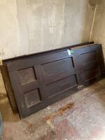 Five solid dark wood interior doors stacked against a basement wall showing wear and weathering.