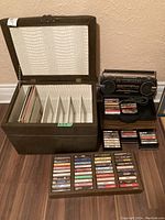 Photo showing open brown faux leather trunk with white crocodile pattern interior and divided compartments, partially filled with vinyl records. Next to trunk is a portable cassette player resting on a black fabric case and several cassette tapes arranged and stacked.