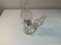 Full view of the clear glass hurricane oil lamp standing on a white surface casting a shadow.