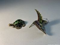 Photo showing the cloisonné turtle and hummingbird ornaments side by side on a plain surface, highlighting the size and enamel detail.