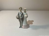 Front view of Lladro porcelain figurine showing bride and groom standing together, groom holding hat, bride holding bouquet.