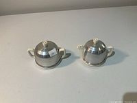 Two pieces of a vintage creamer and sugar bowl set. Both have dome-shaped chrome lids and cream-colored ceramic bodies with handles. Positioned side-by-side on a white surface.