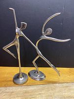 Front view of two chrome metal dancing figurines on oval bases showing abstract human dance poses.
