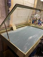 Open view of the vintage glass display case showing interior lined with worn blue fabric and metal frame with hinged lid held open by bracket.