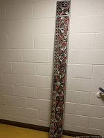 Full length photo of long narrow hand carved and painted wood panel showing overall size and shape leaning against a wall.