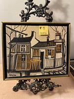 Frontal view of the oil painting in its frame on a decorative stand, showing the full artwork with two buildings and the sign AU LAPIN AGILE.
