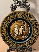 Front view of the antique Majolica Austrian plate displayed on a decorative metal stand showing the raised white figurines of a boy and girl in the center against a glazed background with floral border.