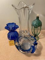 Four pieces of art glass displayed on table including 2 vases, 1 blue and clear ashtray, and 1 green glass lamp