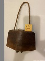 Front view of a large vintage wooden cow bell suspended by a thick braided rope handle against a white background. Shows the carved wood surface and rectangular shape.