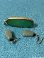 Photo showing green jade pendant with gold-tone trim alongside two green semi-precious stone earrings with gold-tone hooks on a blue surface.