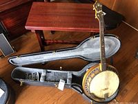 Full view of vintage Humphrey banjo laid in open hard case on wood floor, showing wooden neck, metal rim, and functional hardware
