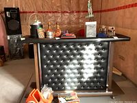 Front view of portable bar showing black vinyl padded front with button tufting, wooden sides, metal trim, and items on top (not included).
