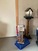 Image showing wooden welcome sign bird feeder on pedestal, small hanging bird feeder on blue plastic basket, and unopened bag of bird feed in background.