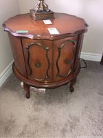 Front angled view of round wooden end table showing scalloped edge top, two-door storage area with decorative hardware, and cabriole legs. Wear visible on wood finish.
