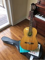 Full view of vintage Kent acoustic guitar leaning on a piano with the black hard case on the floor.