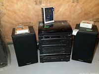 Wide view showing two Magnavox bookshelf speakers flanking a stacked stereo system including turntable, dual cassette, DVD player, and receiver/amplifier. Items are black and in basement setting.