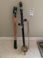Photo showing a decorated didgeridoo, a black wooden rabbaba, and a small bamboo flute against a wall on carpeted floor.