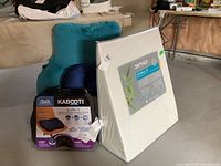 Wide shot showing all items in the lot including the Orthex Symbia 18 posture cushion, Kabooti 3-in-1 back rest pillow in box, a small blue neck pillow, and storage bin.