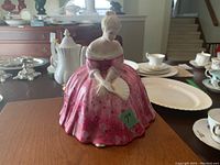 Front view of Royal Doulton Victoria 7 inch porcelain figurine in pink dress holding fan placed on wood surface with various china dishes in background.