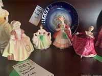 Four Royal Doulton figurines displayed on a shelf, showing three standing female figures in various dresses and one holding flowers.