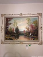Front view of signed landscape painting in ornate cream frame hung on wall showing autumn river scene.