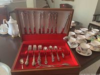 Photo showing opened wood box containing 15 pieces of silverplate flatware including spoons, forks, and knives. The flatware is neatly arranged in slots with a red lining. The box features a handle and storage drawer below.
