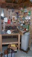 Wide angle photo showing wooden workbench with pegboard of tools including saw, hammer, clamps, and assorted small hardware items in packages and drawers. Hard hat and other hand tools on workbench surface.